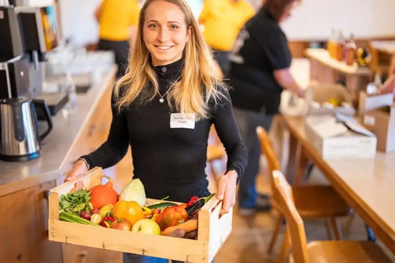 Lena Fischl trägt eine Kiste mit frischem Bio-Gemüse und Obst auf dem Hof